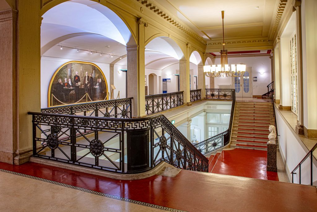 Wandelhalle im LVH Langenbeck Virchow Haus Berlin mit rotem Boden, Treppenaufgang und historischem Gemälde als repräsentative Eventlocation