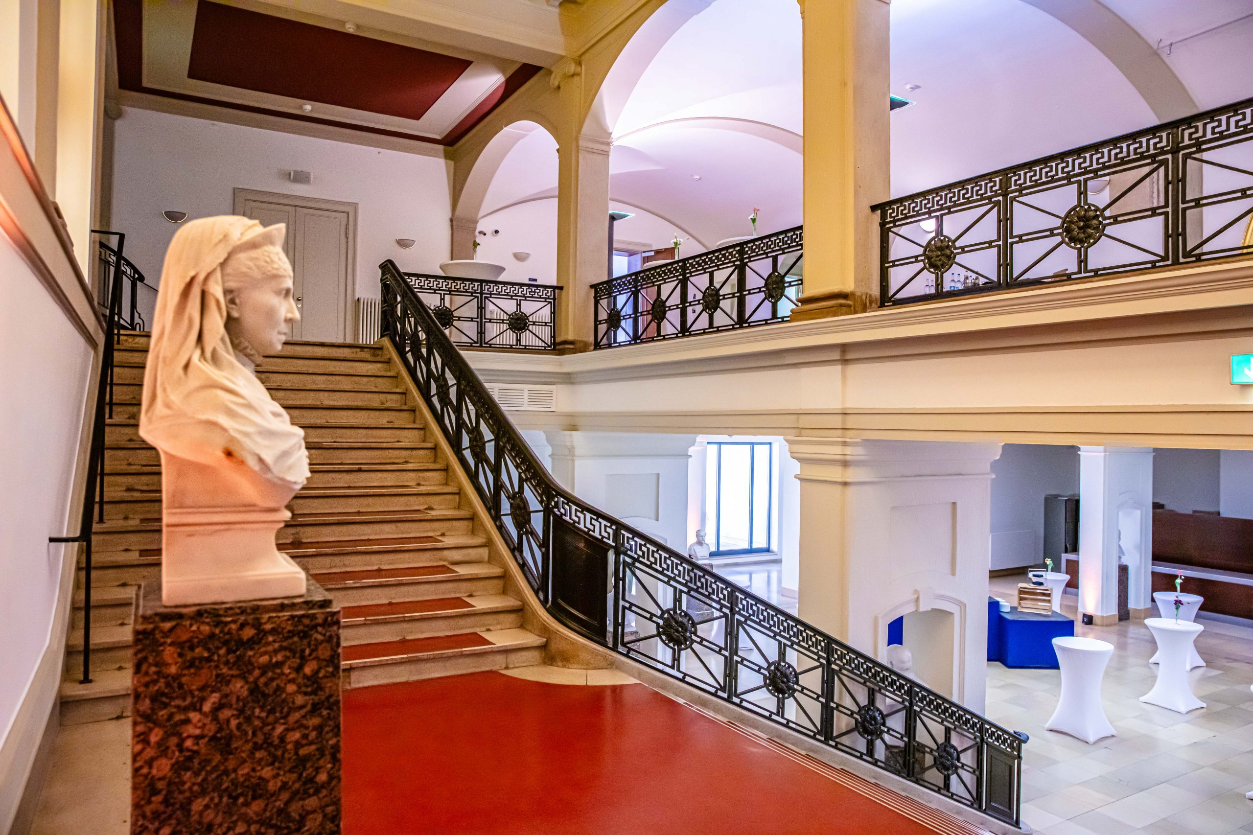 Historisches Foyer im Erdgeschoss des LVH Langenbeck Virchow Haus Berlin mit rotem Boden, Treppenaufgang und Skulptur als repräsentative Eventlocation